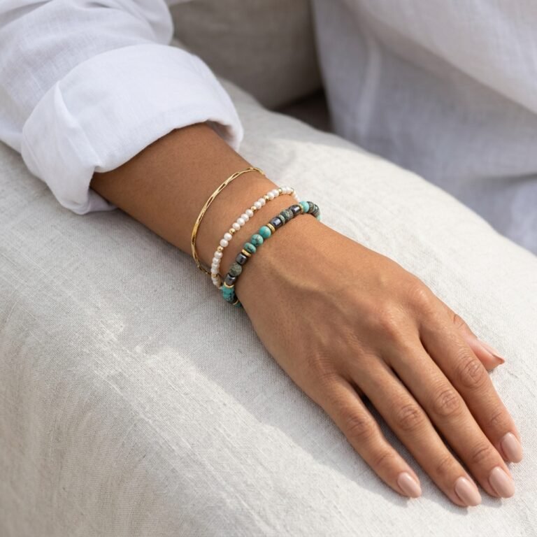 Close-up do pulso de uma mulher usando uma combinação de três pulseiras: um bracelete rígido dourado, uma pulseira delicada de pequenas pérolas brancas e uma pulseira de contas de pedras turquesa e cinza-escura. Ela veste uma camisa de linho branca e apoia a mão com unhas pintadas de nude sobre um tecido claro.